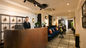 A reception area with a receptionist, a reception desk labeled "Carillon Reception," a blue sofa, patterned floor tiles, and a staircase in the background. - Carillon Boutique Hotel (Hanoi)