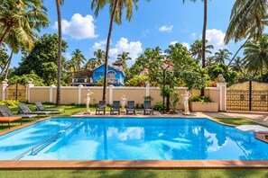 A swimming pool with lounge chairs and statues, surrounded by palm trees and a blue house in the background. - Villa Calangute Phase 4 (Calangute)