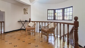 Ein geräumiges Zimmer mit Holzboden, zwei Korbstühle mit Kissen, einem kleinen Tisch und einem großen Fenster mit Buntglas.