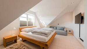 A bedroom with a sloped ceiling, a bed with pillows, a nightstand, a sofa, a TV mounted on the wall, and a window with blinds.