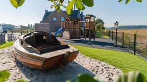 A wooden boat in a sandy area with a playground and a house in the background.