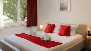A neatly made bed with red and white bedding, a red throw blanket, and two towels. There is a small bedside table with a lamp and a framed picture on the wall. - Grillon by Interhome (Zermatt)