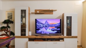A modern living room with a flat-screen TV on a wooden entertainment unit, a dining table with chairs, and a potted plant. - Grillon by Interhome (Zermatt)