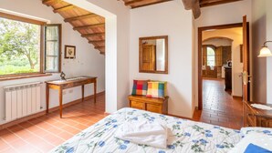 A bedroom with a sloped ceiling, a bed with a floral duvet, a wooden dresser, a mirror, and a window with a view of greenery. - Baccelli by Interhome (Montaione)