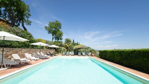 A rectangular swimming pool with clear blue water, surrounded by white lounge chairs and umbrellas, set against a backdrop of trees and a stone wall. - Baccelli by Interhome (Montaione)