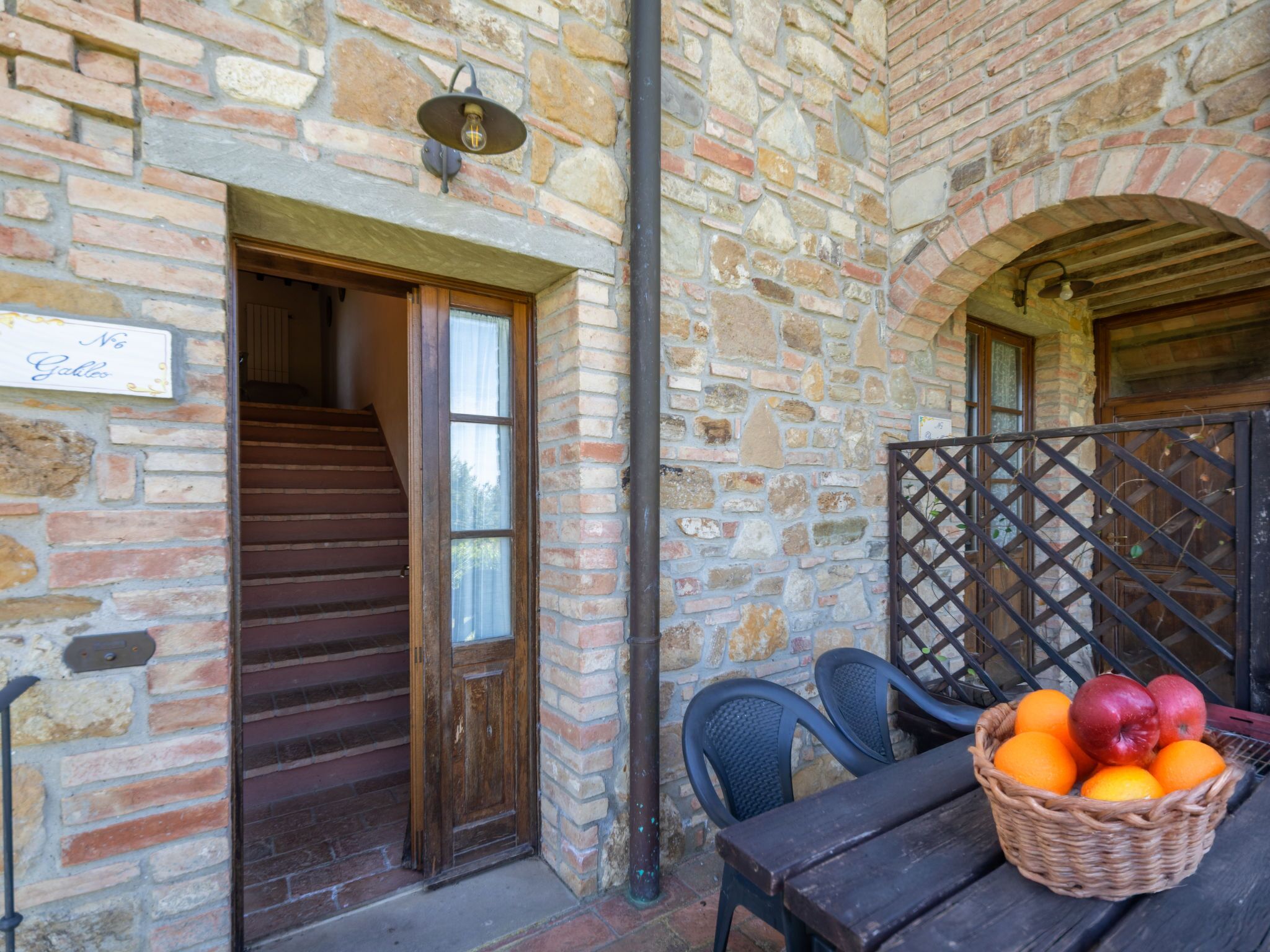 A stone building with a wooden staircase leading to an open door, a wicker basket of fruit on a table, and a black metal gate.