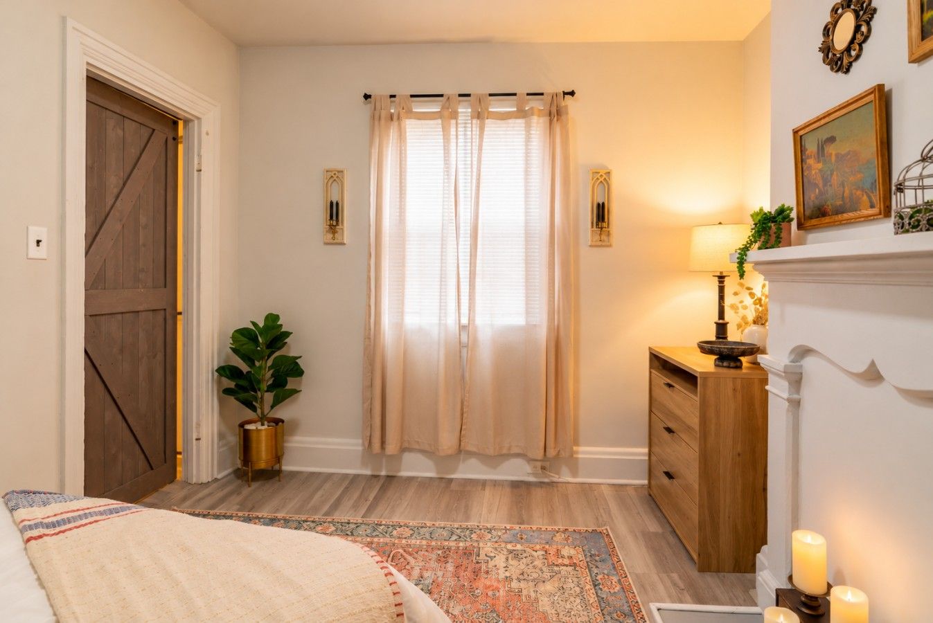 A bedroom with a wooden door, a large window with curtains, a bedside table with a lamp, a potted plant, and a patterned rug.