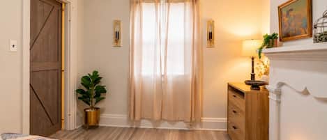 Un dormitorio con puerta de madera, un ventanal con cortinas, una mesita de noche con lámpara, una planta en maceta y una alfombra estampada.