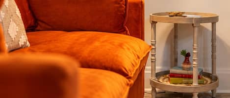 A room with a sofa, a lamp, a small table with books, and a framed artwork on the wall.