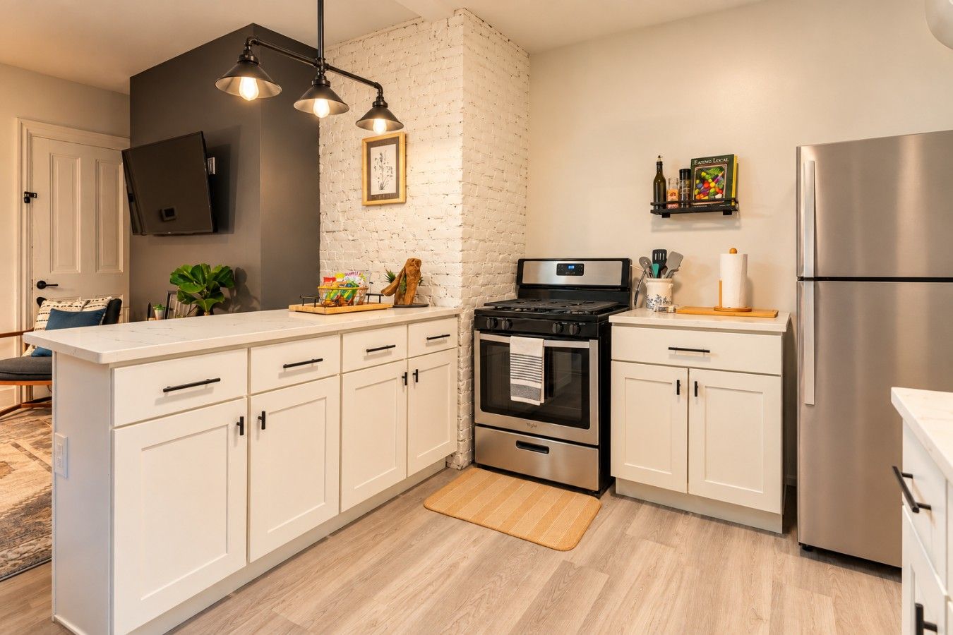 A modern kitchen with white cabinets, a stainless steel refrigerator, a stove, and a flat-screen TV mounted on the wall.
