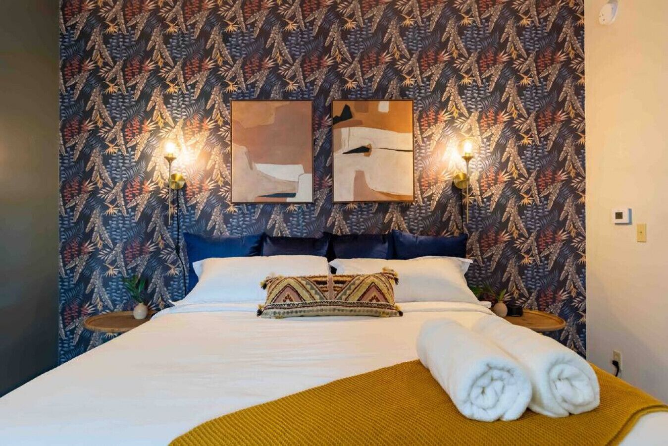 Een bed met witte lakens, een gestreept plaid en een opgerolde handdoek. Er zijn twee wandlampen en twee ingelijste kunstwerken boven het bed.