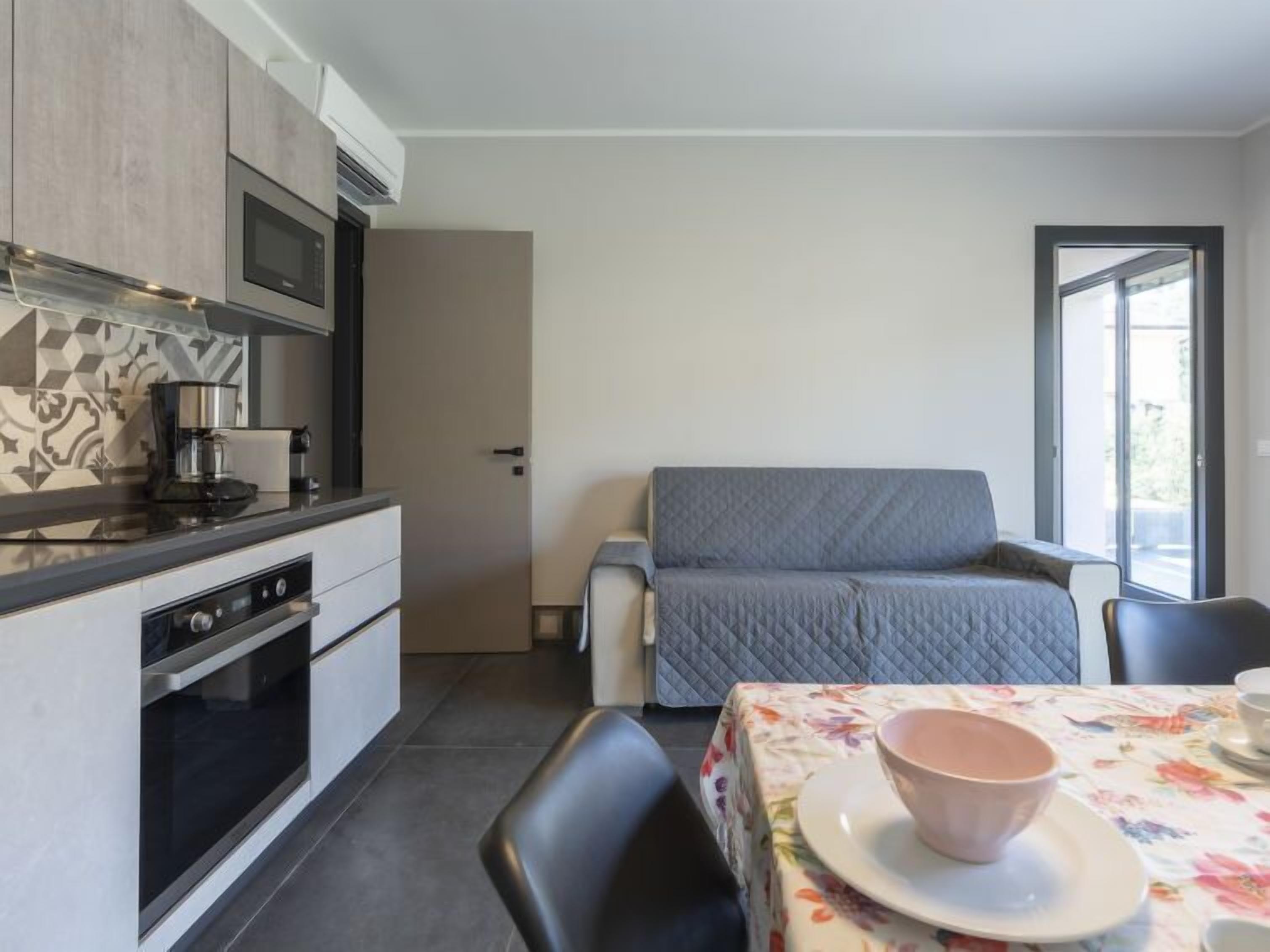 Uma cozinha compacta com eletrodomésticos modernos, uma área de refeições com toalha de mesa florida e um sofá cinzento.