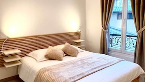 Una cama bien hecha con cabecera de madera, una mesita de noche con lámpara y una ventana con cortinas.