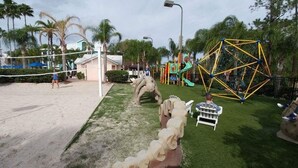 A beach volleyball court with a net, palm trees, and a playground with a climbing structure and slides.