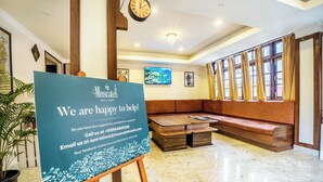 A hotel lobby with a reception desk, a clock, a large window with curtains, a seating area, and a sign offering help. - Muscatel Dzeemkhang (Gangtok)