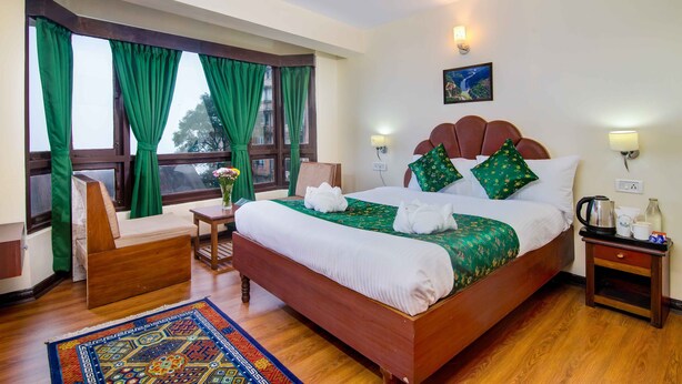 A hotel room with a large bed, green curtains, a wooden chair, a small table, and a nightstand with a kettle and cups. - Muscatel Dzeemkhang (Gangtok)