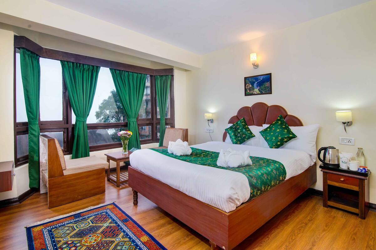 Ein Hotelzimmer mit einem großen Bett, grünen Vorhängen, einem Holzstuhl, einem kleinen Tisch und einem Nachttisch mit Wasserkocher und Tassen.
