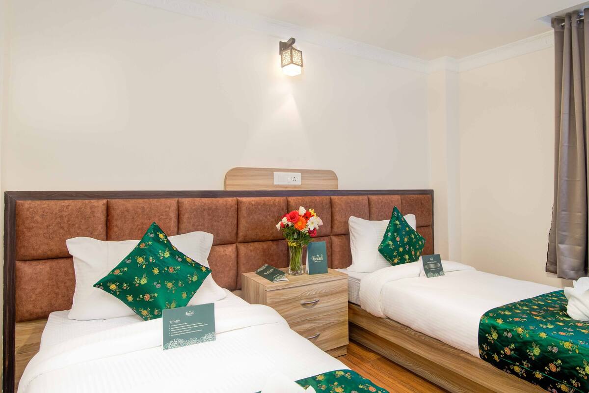 Ein Hotelzimmer mit zwei Betten, jeweils mit weißer Bettwäsche und grün gestreiften Kissen. Zwischen den Betten befindet sich ein Nachttisch mit Blumen und einer Karte.