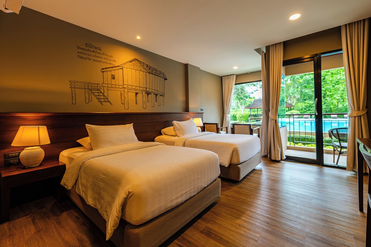 A hotel room with two beds, a nightstand, a lamp, a wall mural, and a view of a swimming pool.