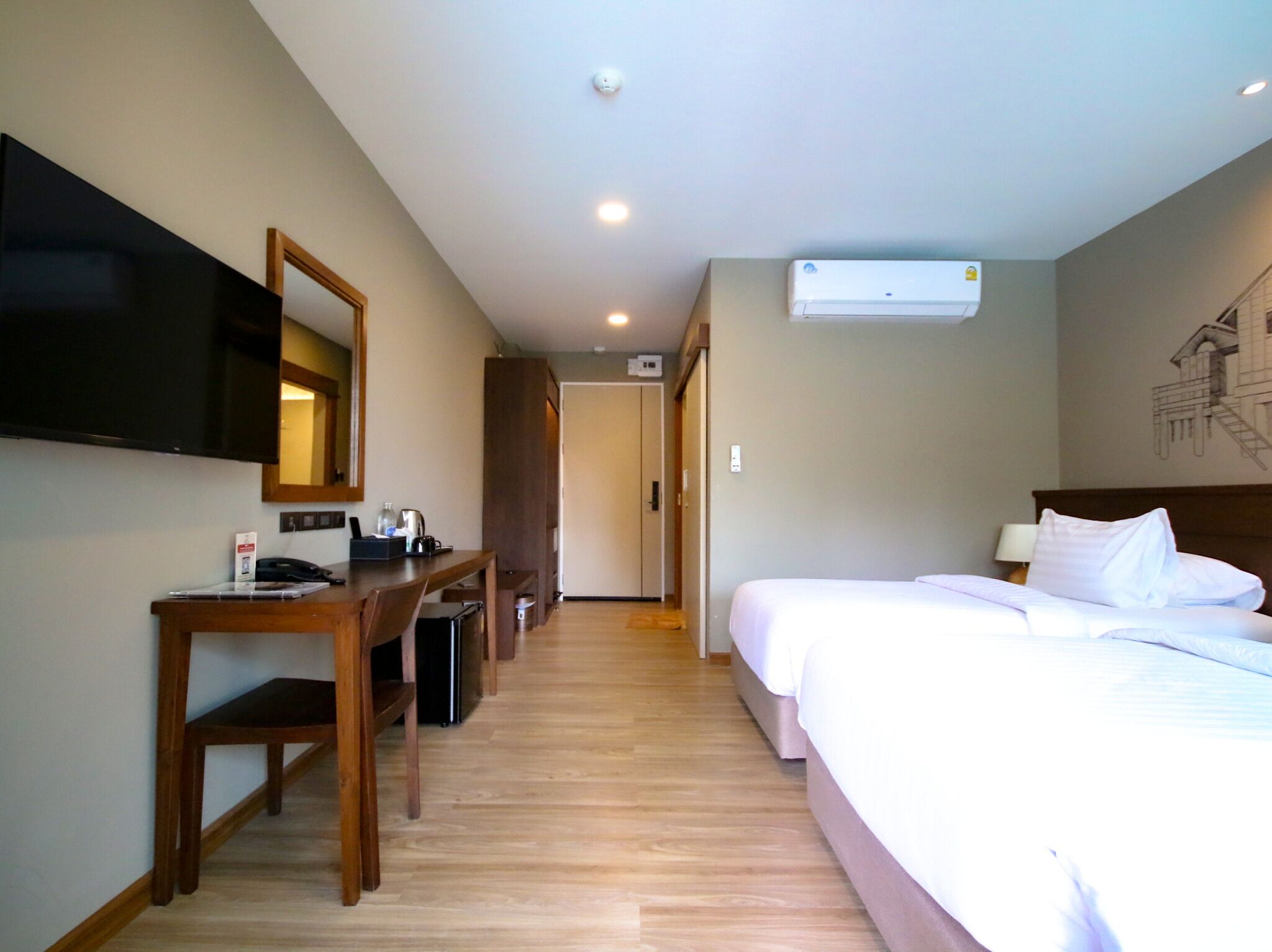 A hotel room with a bed, a desk with a chair, a flat-screen TV mounted on the wall, and an air conditioning unit.