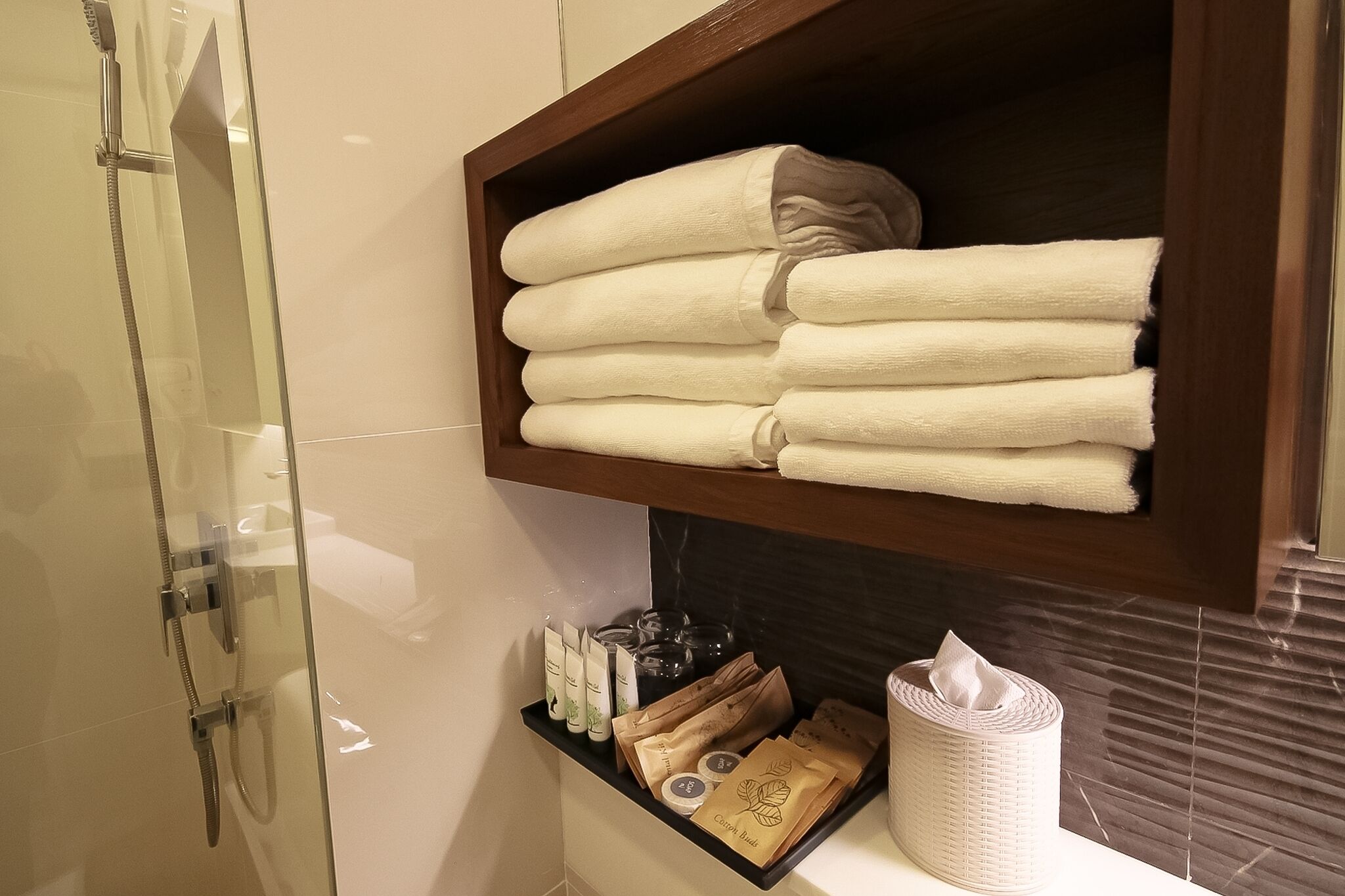 A bathroom with a glass shower enclosure, a wooden shelf with folded towels, toiletries, and a tissue roll.
