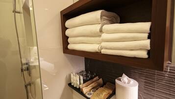 A bathroom with a glass shower enclosure, a wooden shelf with folded towels, toiletries, and a tissue roll.