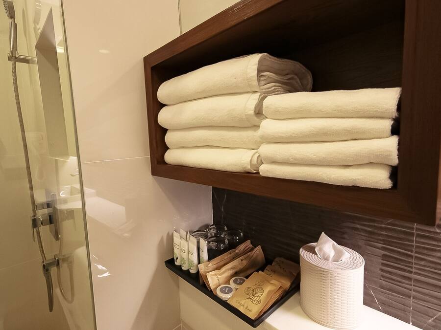 A bathroom with a glass shower enclosure, a wooden shelf with folded towels, toiletries, and a tissue roll.