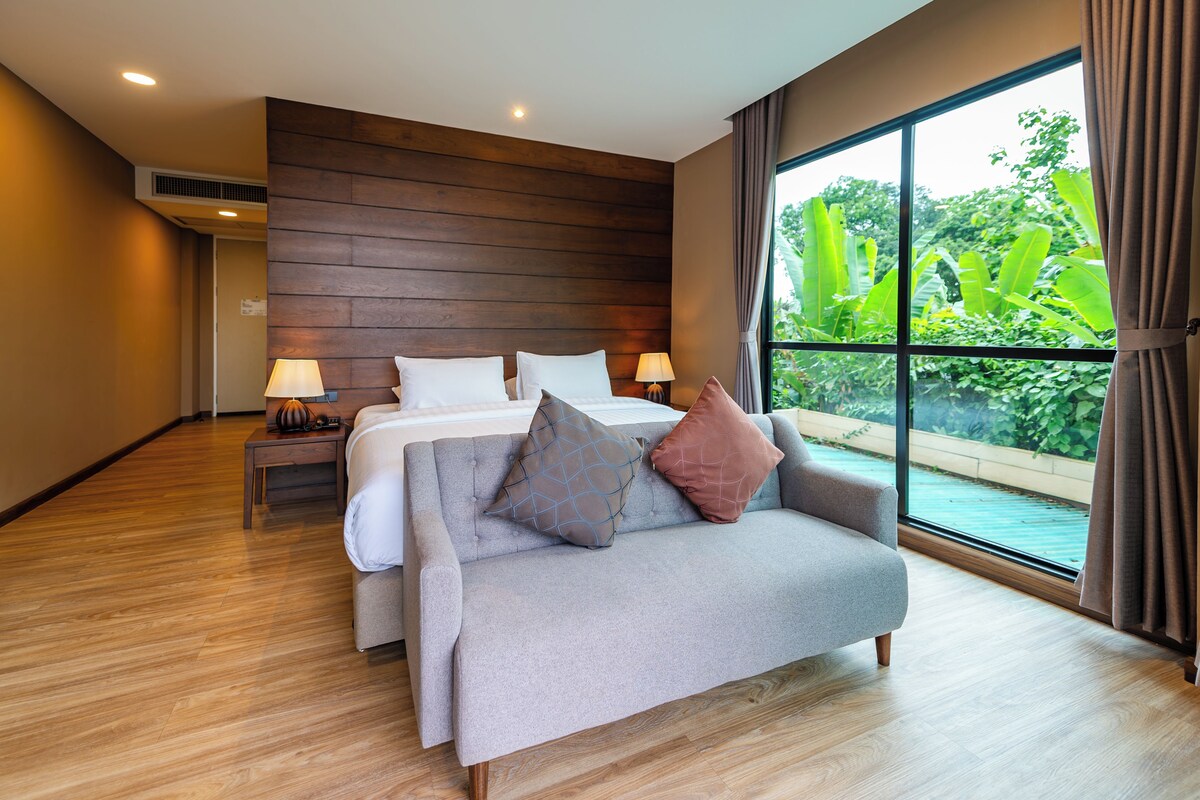 A modern hotel room with a large bed, a sofa, two bedside tables with lamps, and a view of greenery through a large window.