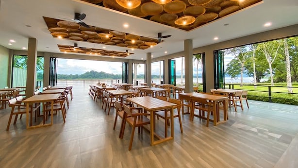 A spacious dining area with wooden tables and chairs, large windows offering a view of the outdoors, and a ceiling adorned with woven light fixtures. - RiverTree Resort Chiangkhan  (Chiang Khan)