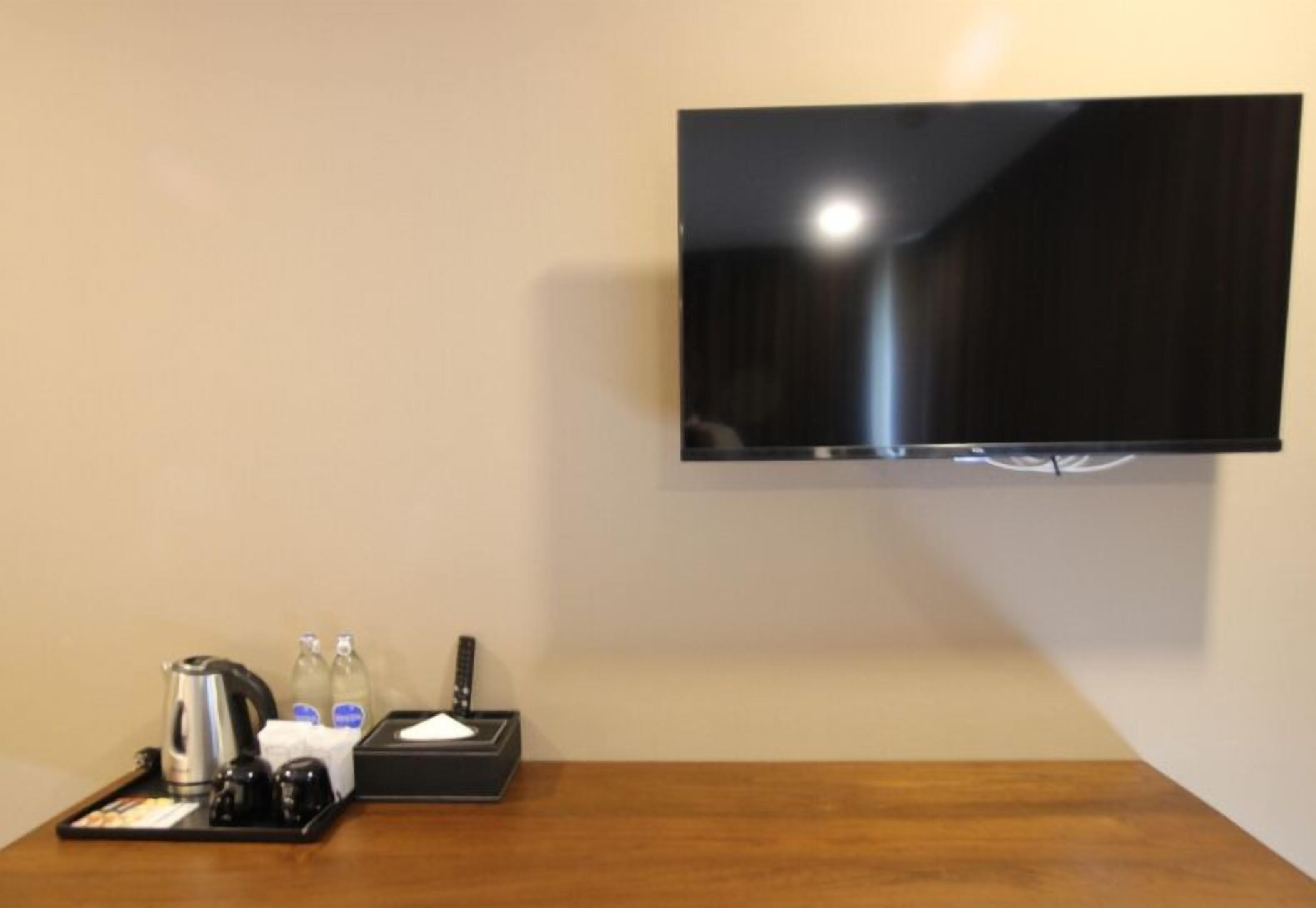 Habitación de hotel con una televisión de pantalla plana montada en la pared, una mesa de madera con una bandeja que contiene una tetera, botellas y un control remoto.