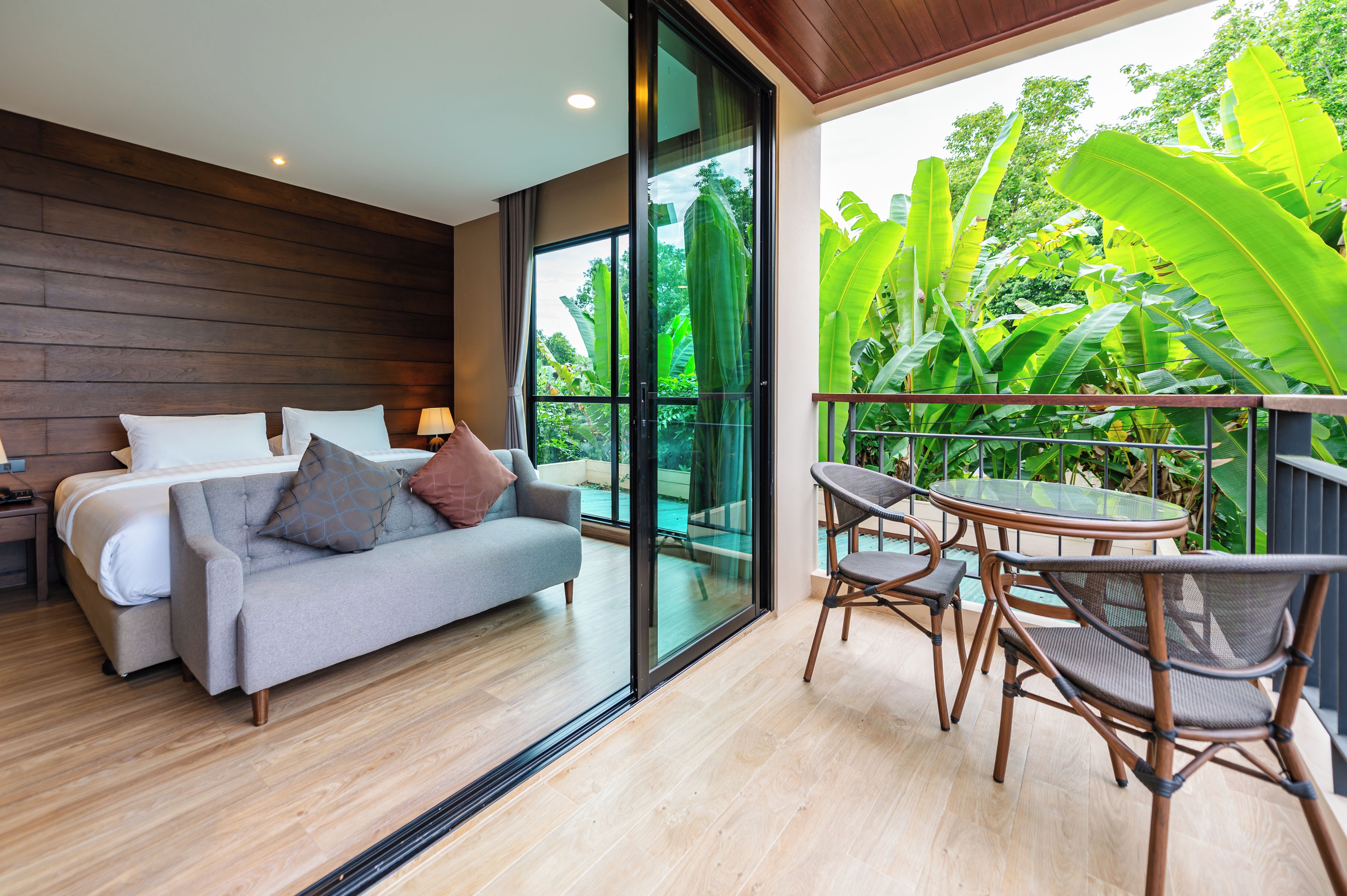A modern bedroom with a sofa bed, a small table, and chairs, a large window with a view of greenery, and a balcony with a glass table.