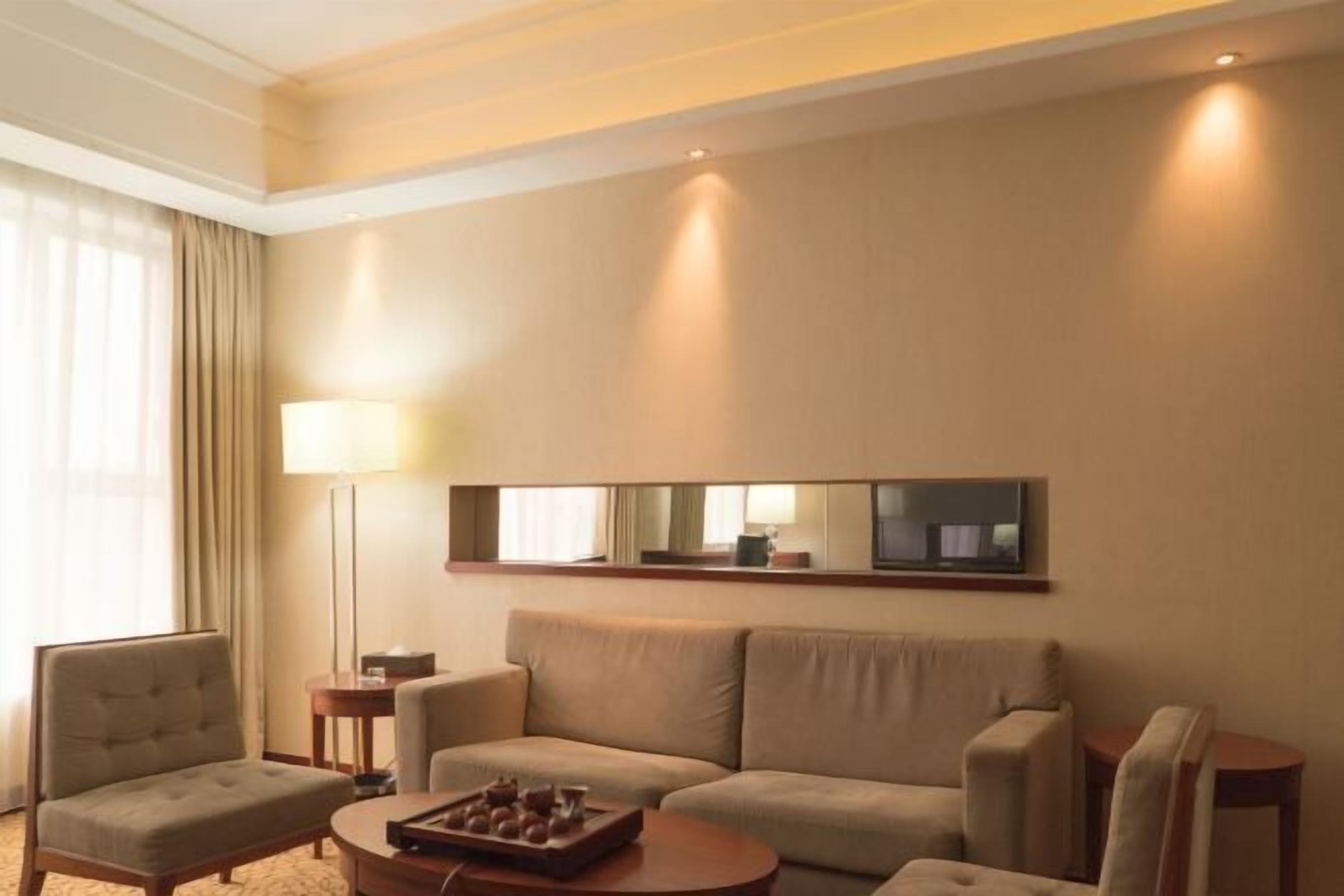 Habitación de hotel con sofá, sillones, una mesita de centro, lámpara y televisor de pantalla plana montado en la pared.