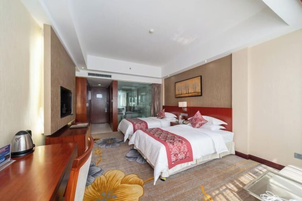 A hotel room with two beds, a TV, a desk with a kettle, and a glass partition.
