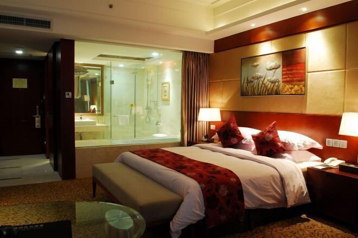 A hotel room with a bed, a bedside table, a lamp, a mirror, and a bathroom visible through a glass door.