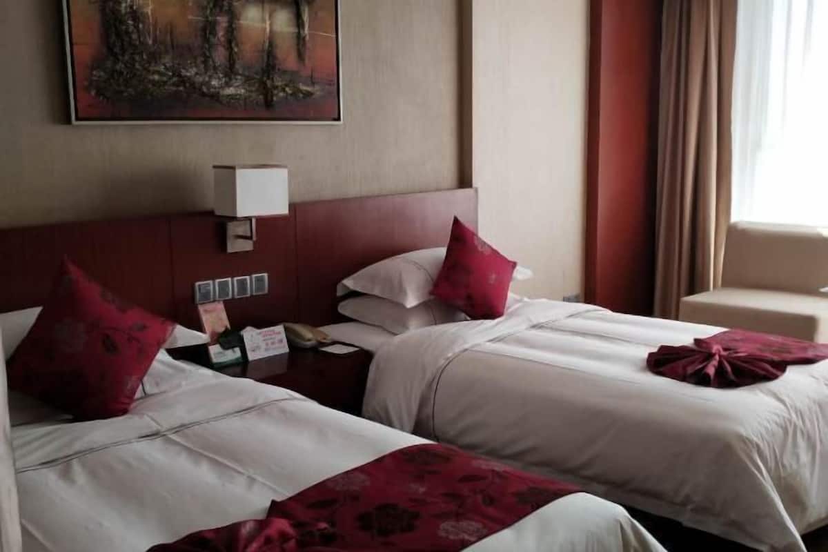 Une chambre d’hôtel avec deux lits, une table de chevet avec une lampe, un tableau au mur et une fenêtre avec des rideaux.