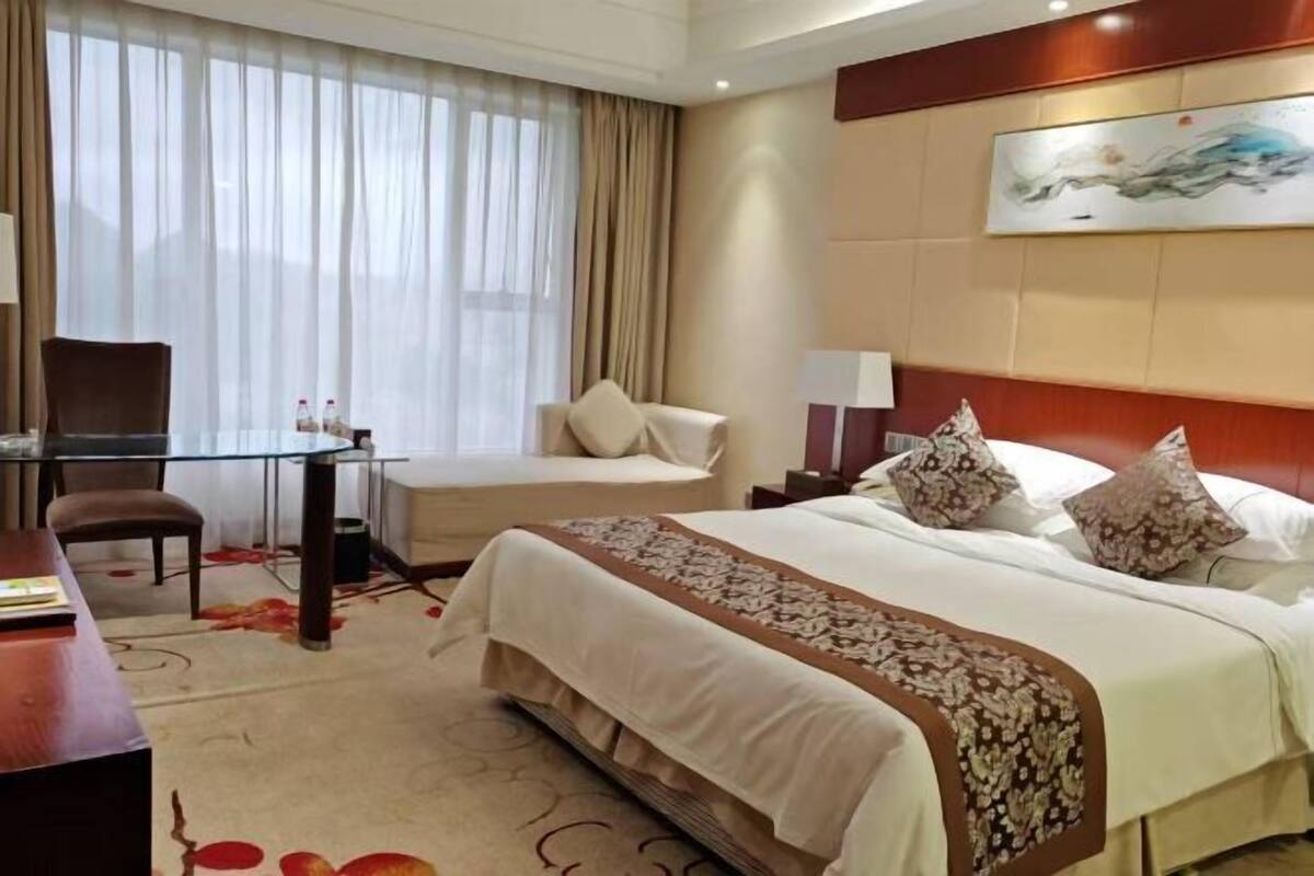 Une chambre d’hôtel avec un lit, un bureau, une chaise, une table et une grande fenêtre avec des rideaux.