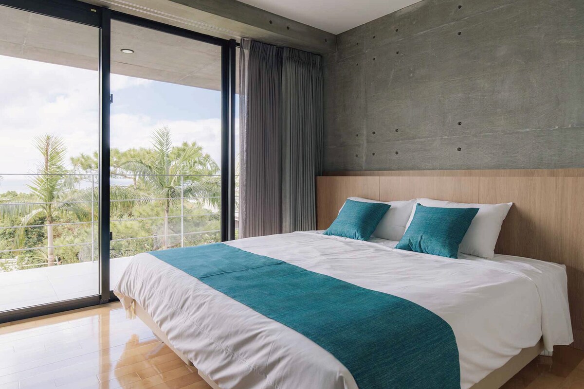 A modern hotel room with a large bed, a concrete accent wall, and a balcony with a view of palm trees.