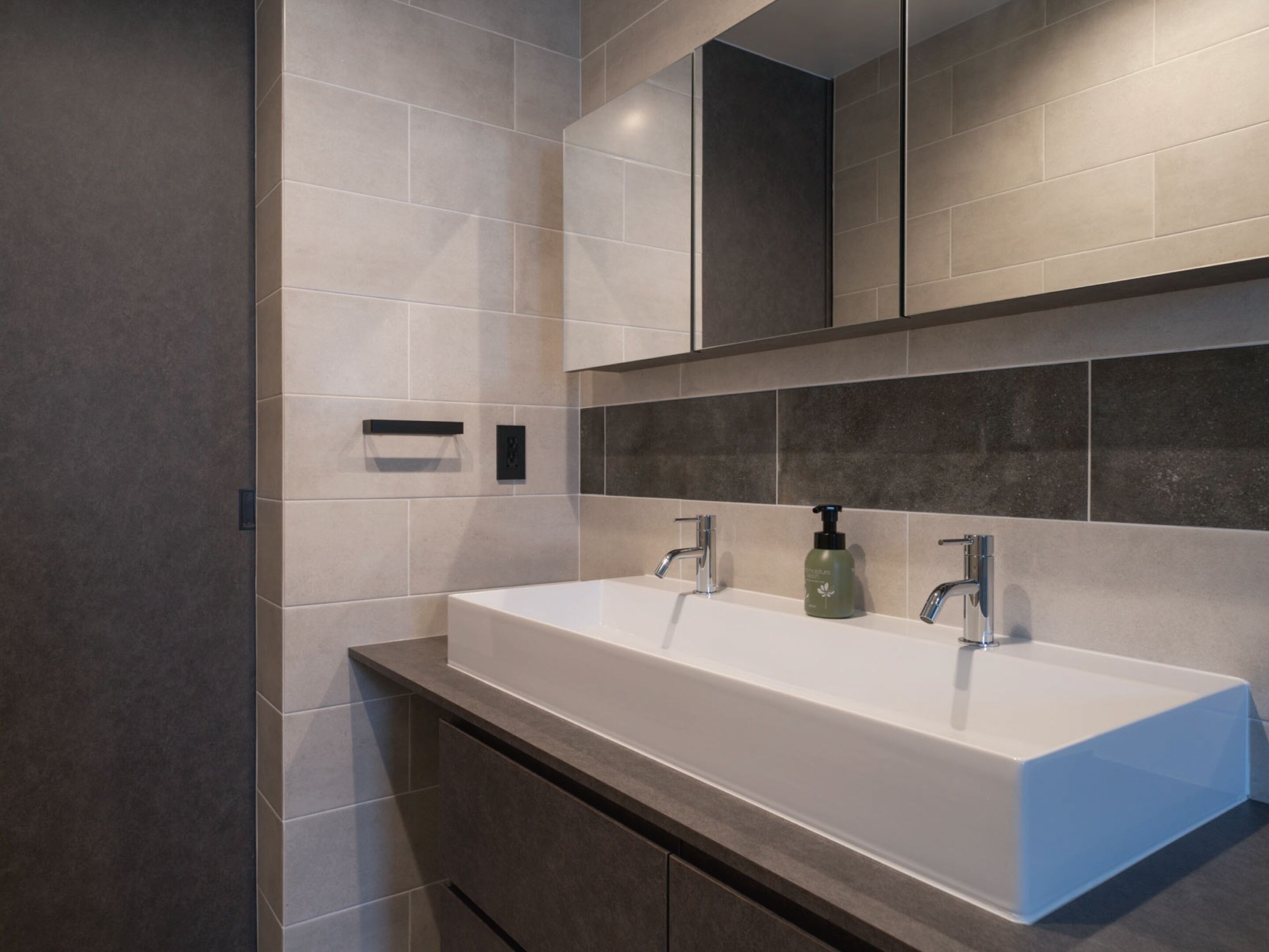 A modern bathroom with a large white sink, dark tiles, and a mirror above.