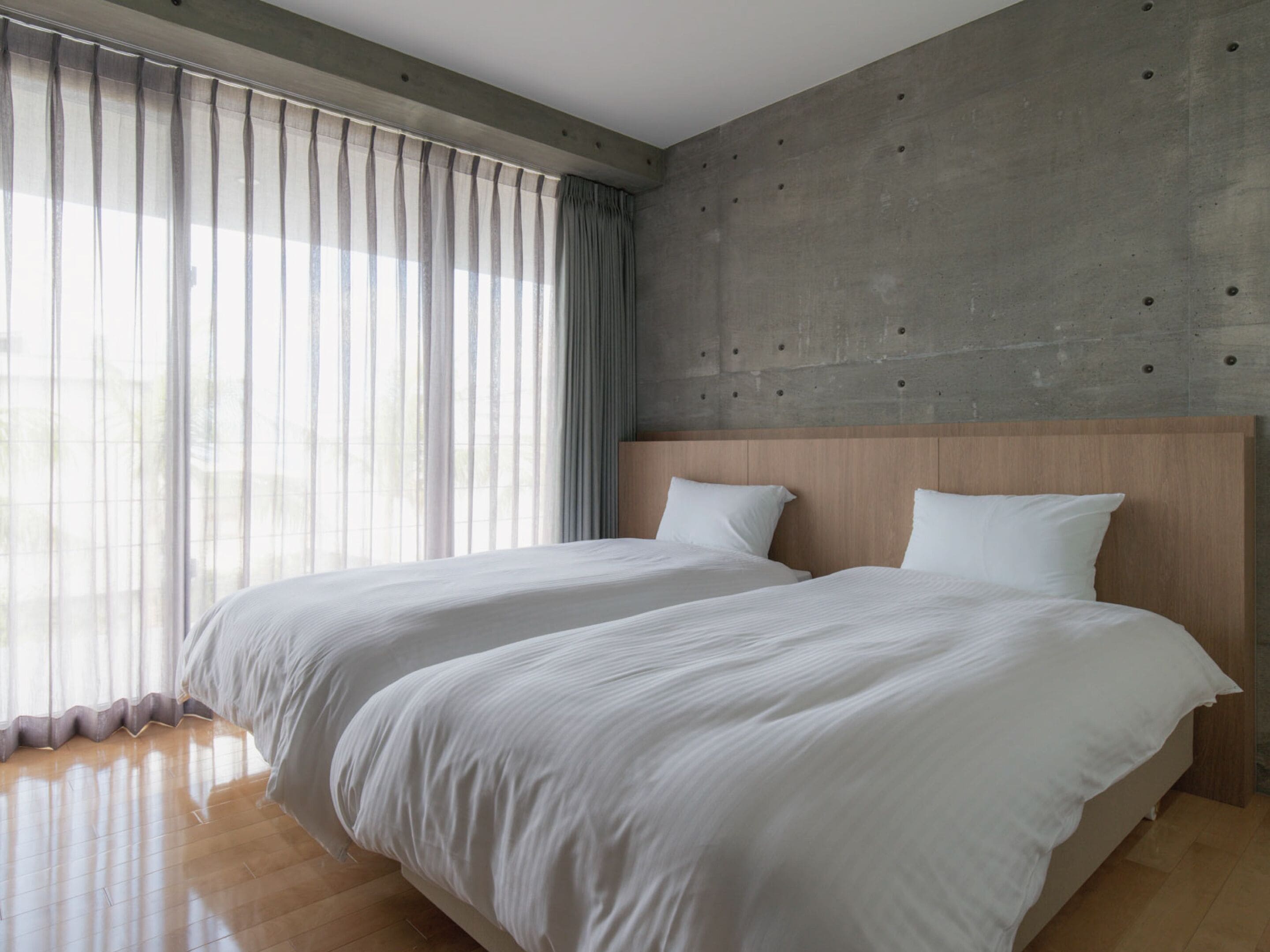 Una habitación de hotel moderna con dos camas, una pared de acento de concreto y ventanas amplias con cortinas traslúcidas.