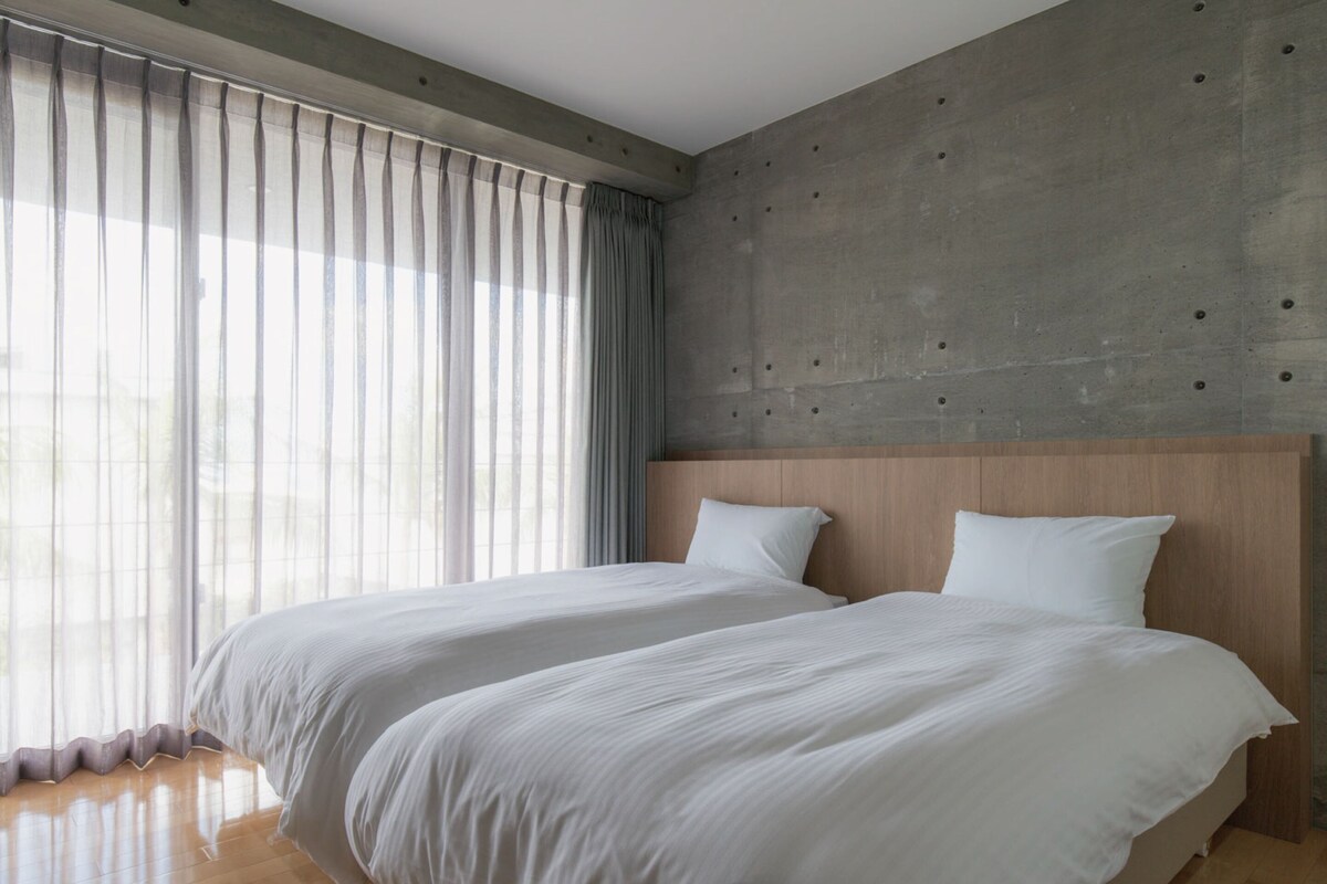 A modern hotel room with two beds, a concrete accent wall, and large windows with sheer curtains.