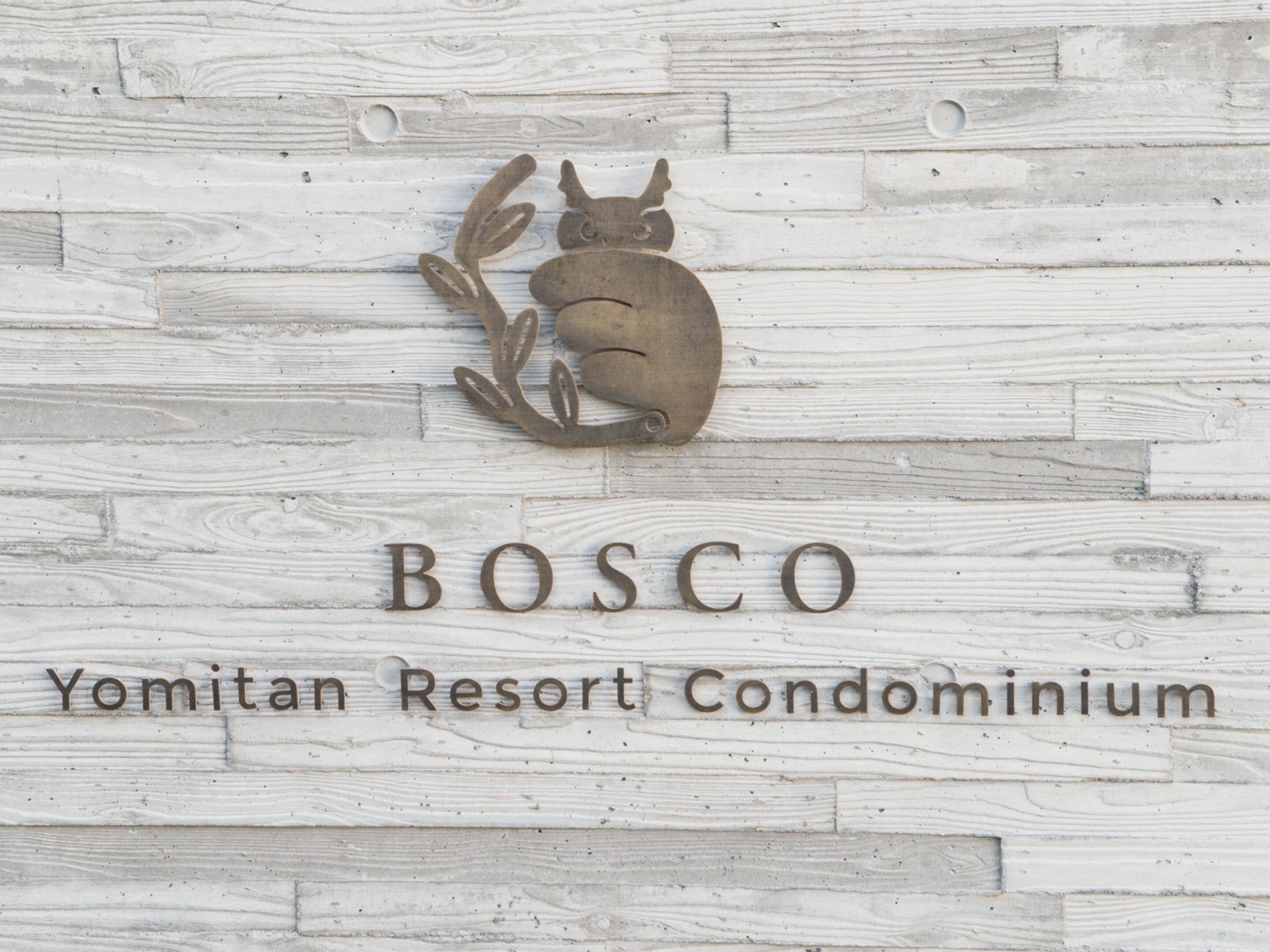 A wall with a wooden texture and a metal sign that reads "BOSCO Yomitan Resort Condominium".
