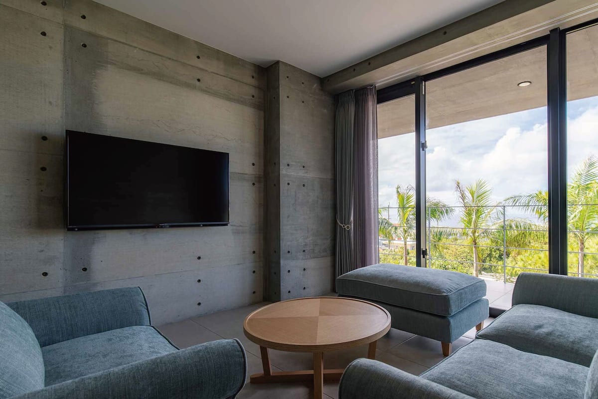 A modern living room with a flat-screen TV mounted on a concrete wall, a round wooden coffee table, and two grey upholstered sofas.