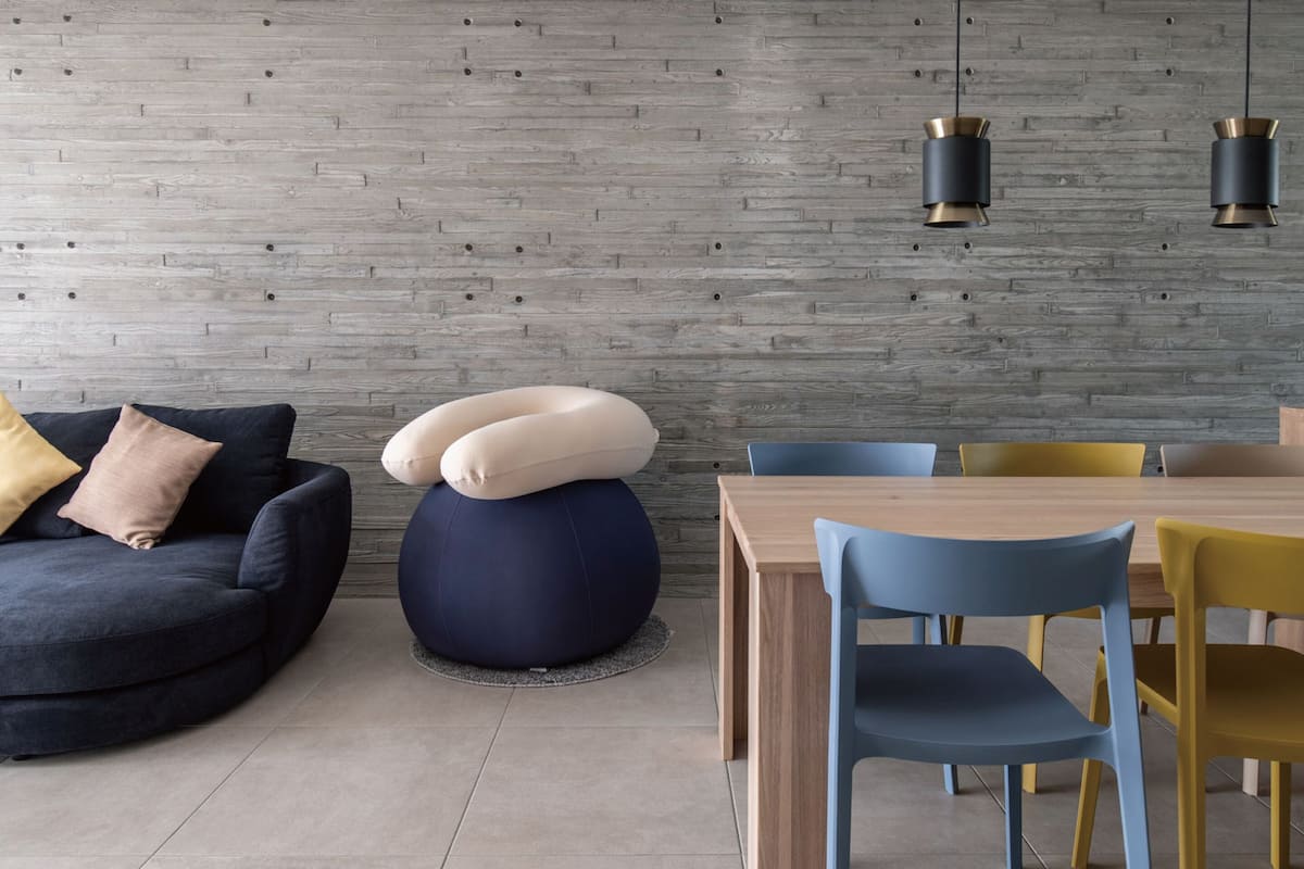 A modern living room with a dark blue sofa, a round blue ottoman, and a wooden dining table with blue and yellow chairs.