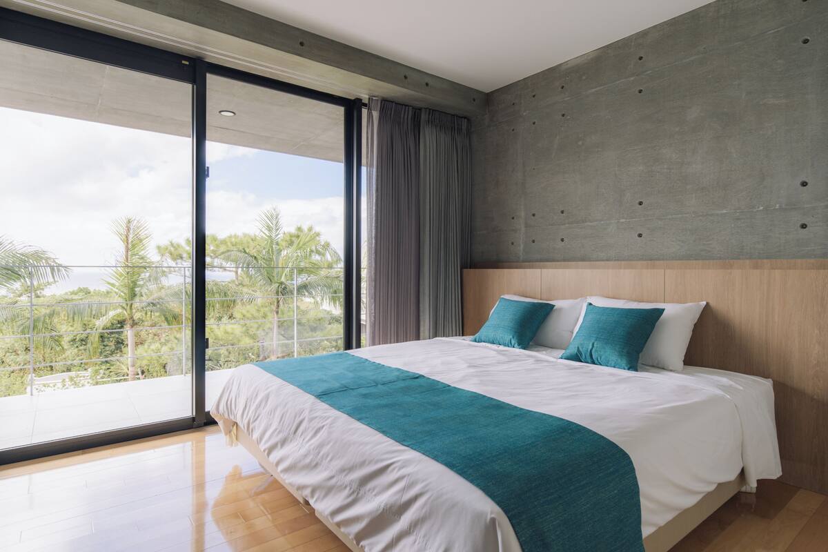 A modern hotel room with a large bed, a view through a sliding glass door, and a concrete accent wall.