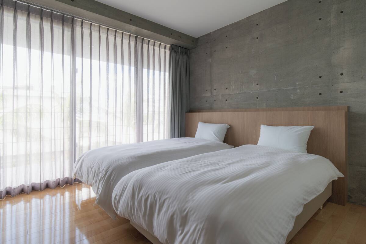 A modern bedroom with two beds, a concrete accent wall, and sheer curtains.