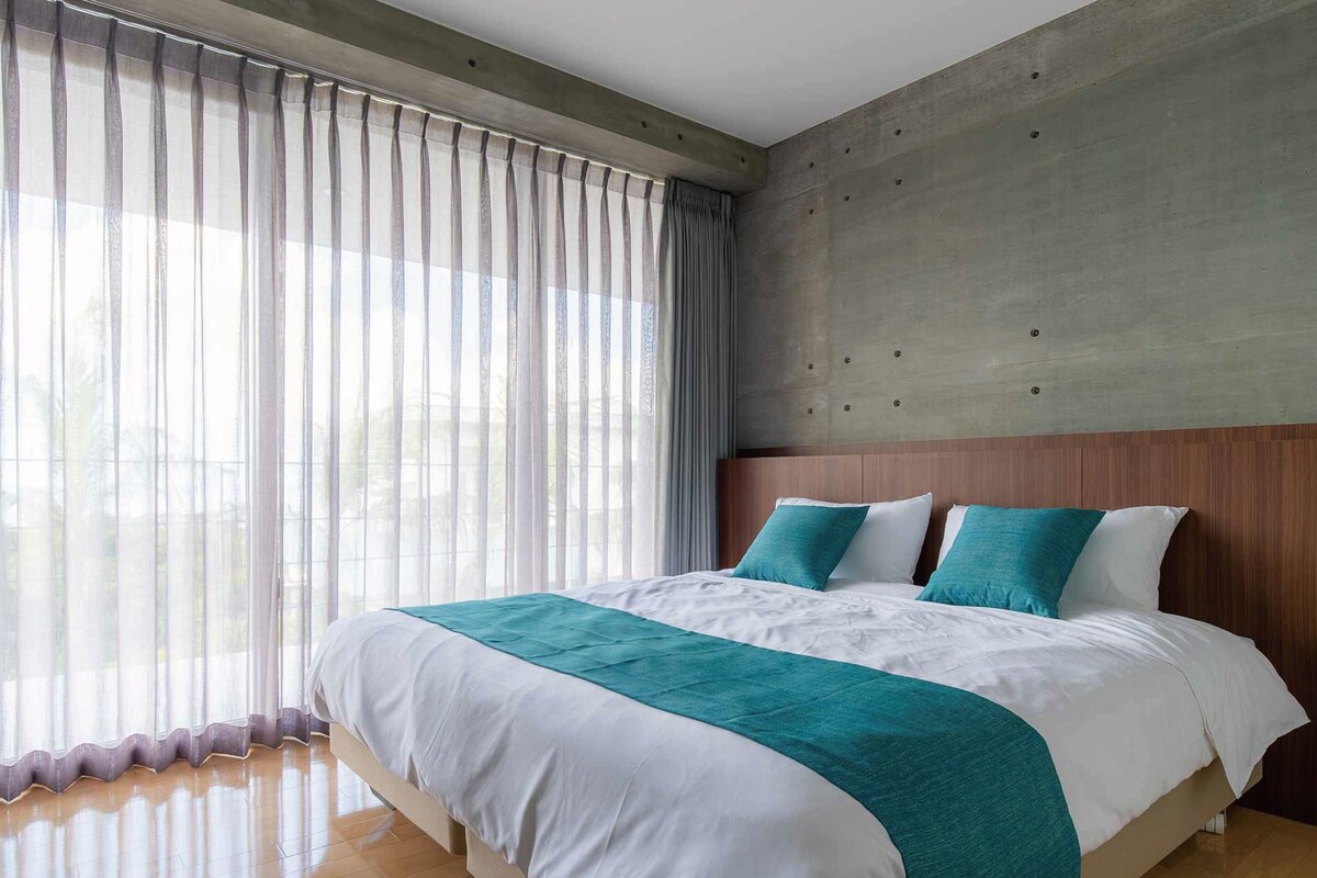 A modern hotel room with a large bed, a concrete accent wall, and sheer curtains.