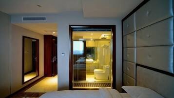 A hotel room with a bed, a large mirror, a sliding door leading to a bathroom, and a view of the city.