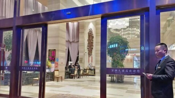 L'entrée d'un hôtel avec un comptoir d'accueil, un espace salon avec des personnes assises, et une enseigne indiquant le nom de l'hôtel.