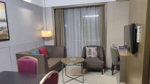Une chambre d’hôtel compacte comprenant un canapé, une petite table ronde, une chaise, un téléviseur et une fenêtre avec des rideaux.