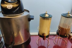 Una cafetera con tapa negra y cuerpo de acero inoxidable, una jarra de vidrio con líquido dorado y un recipiente de vidrio más pequeño con líquido marrón.